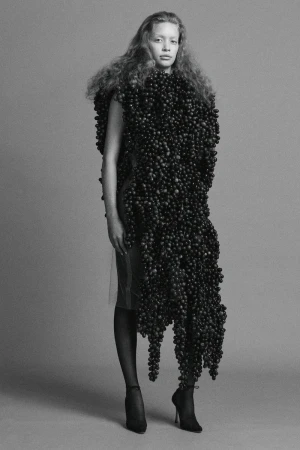 Model wearing an avant-garde sculptural grape garment with sheer tights and heels against a seamless studio backdrop.