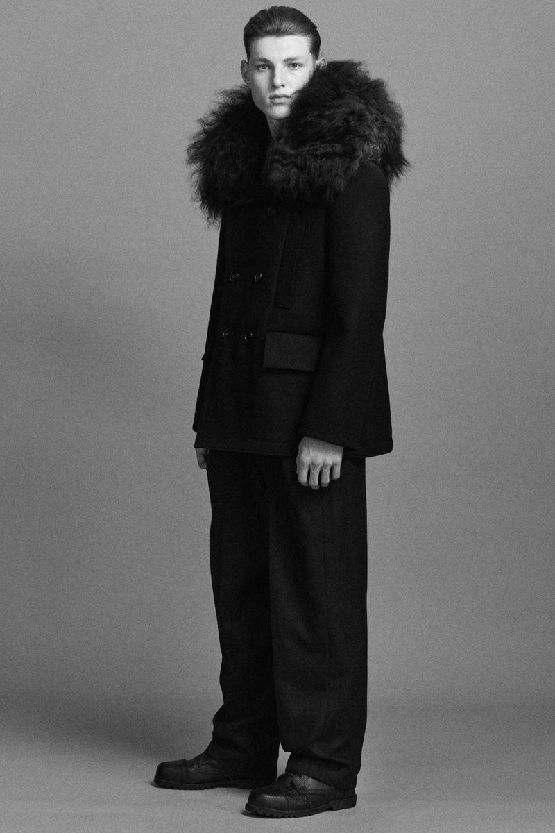 Monochrome studio portrait of a young adult male posing in a dark peacoat with a fur collar, paired with dark pants and dark shoes.