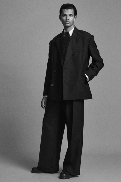 Full body shot of an adult man in a double-breasted black suit with wide-legged pants against a grey backdrop in a studio setting.