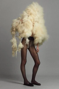 A model standing in a voluminous cream fleece headpiece and top with sheer black tights and black flats against a grey background.