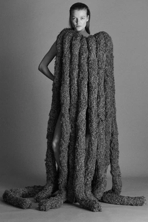 Model wearing an oversized, sculptural grey textured gown with floor-length ropelike strands against a seamless grey backdrop.