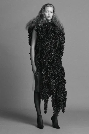 Model wearing an avant-garde sculptural grape garment with sheer tights and heels against a seamless studio backdrop.
