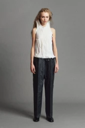 Model Ella Dalton in a crinkled white sleeveless top and dark tailored trousers posing against a grey studio backdrop.