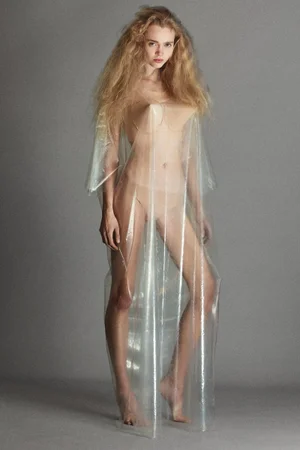 Model with voluminous blonde hair standing in sheer translucent plastic garment against a solid grey studio background.