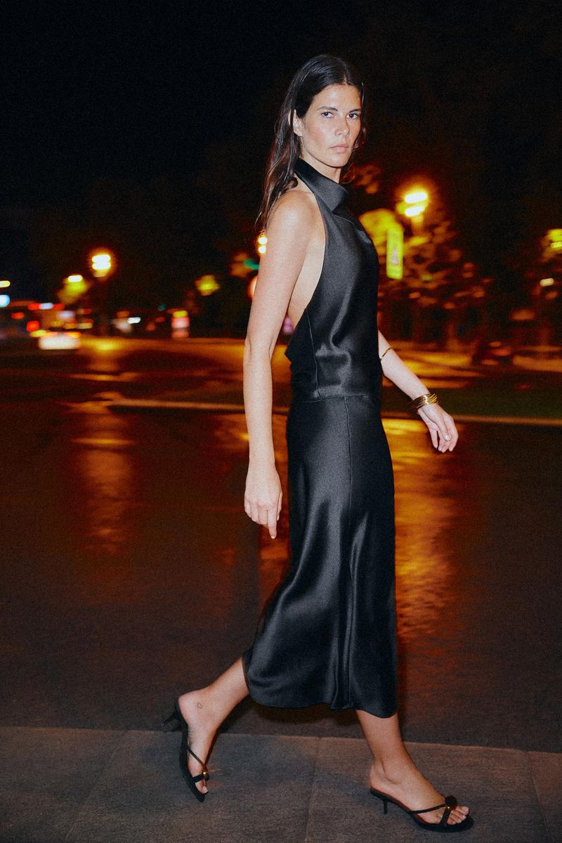 Elegant woman in black halter dress and kitten heels walks on sidewalk at night with city lights reflecting off wet street surface.