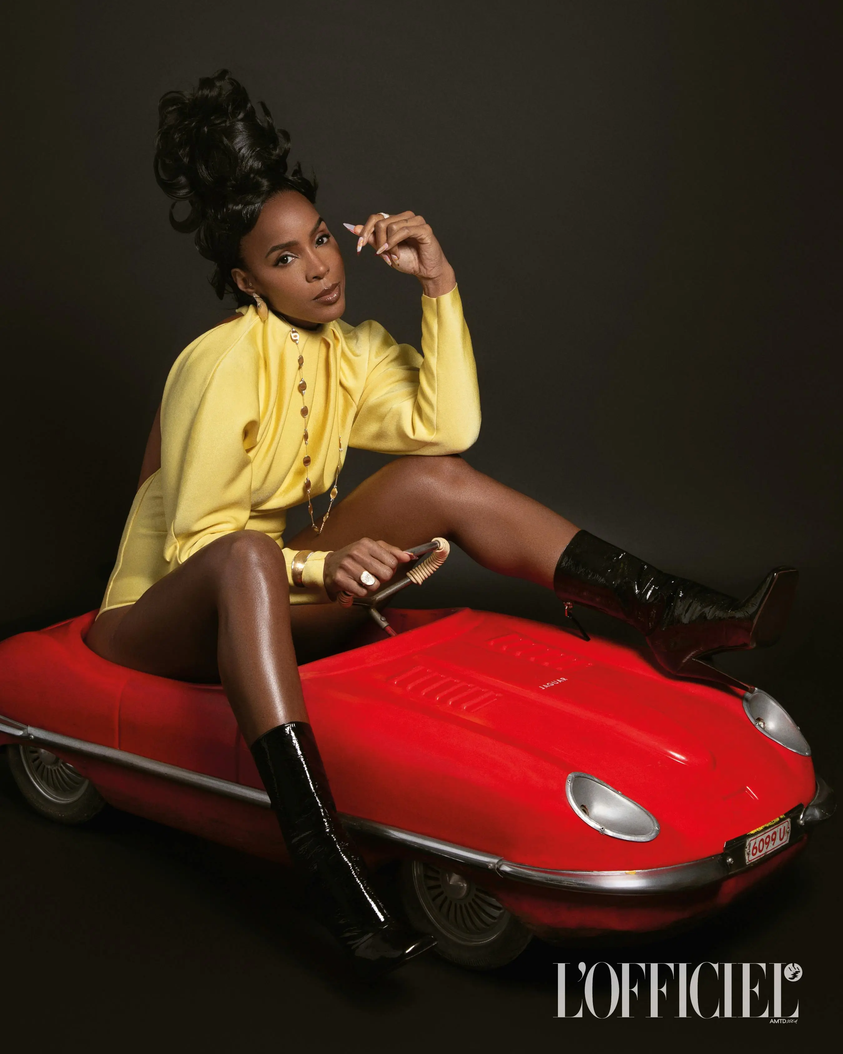 Elegant Black woman poses in a yellow dress atop a red toy car against a dark backdrop, showcasing high fashion and creative set design.
