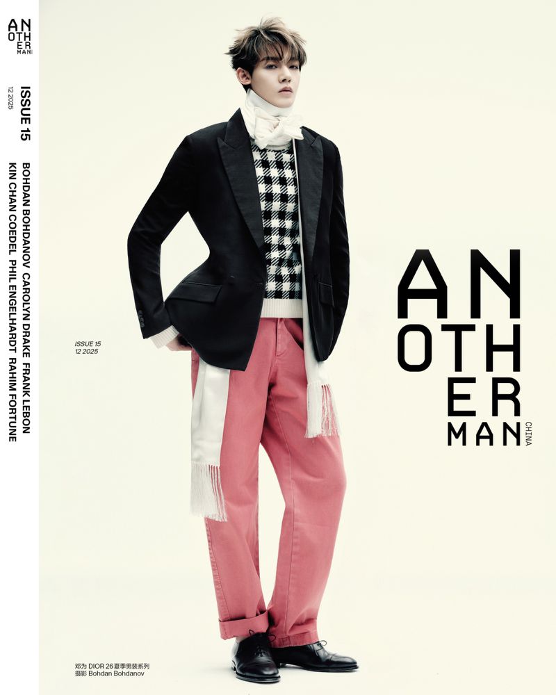 Fashion editorial shot of a male model in a black blazer, patterned sweater, pink pants, and black shoes on a light background.