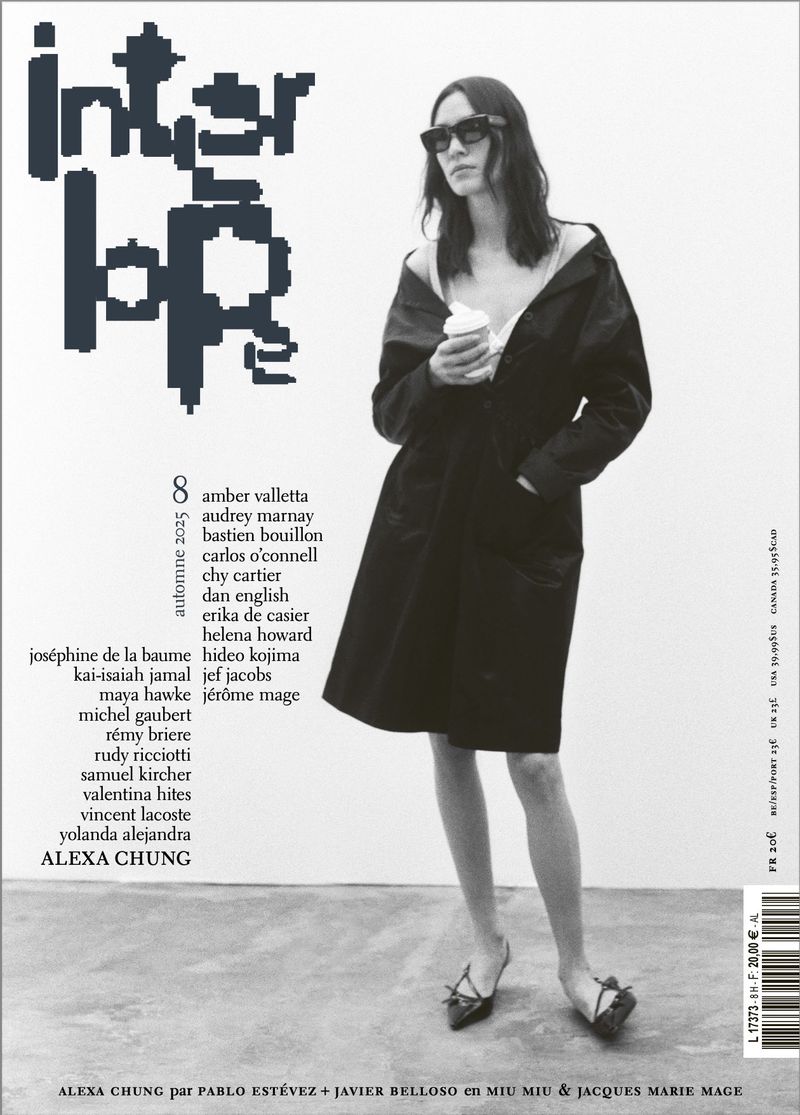 Black and white magazine cover shot of a female fashion model in an oversized black coat and sunglasses, holding a disposable cup.