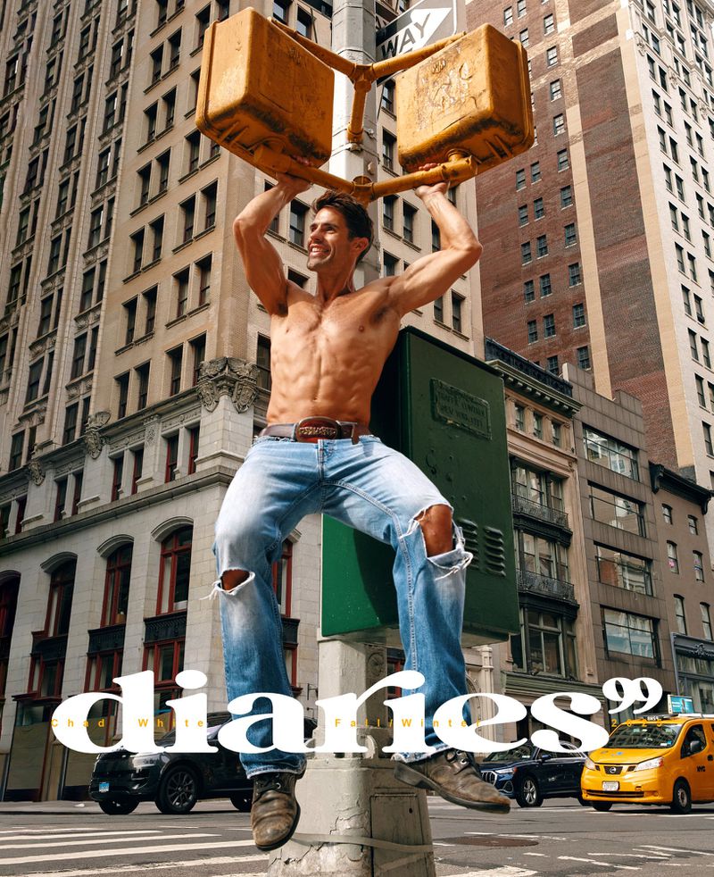 Shirtless muscular man in ripped jeans poses on a city traffic signal, New York. Buildings and city traffic form the urban background.