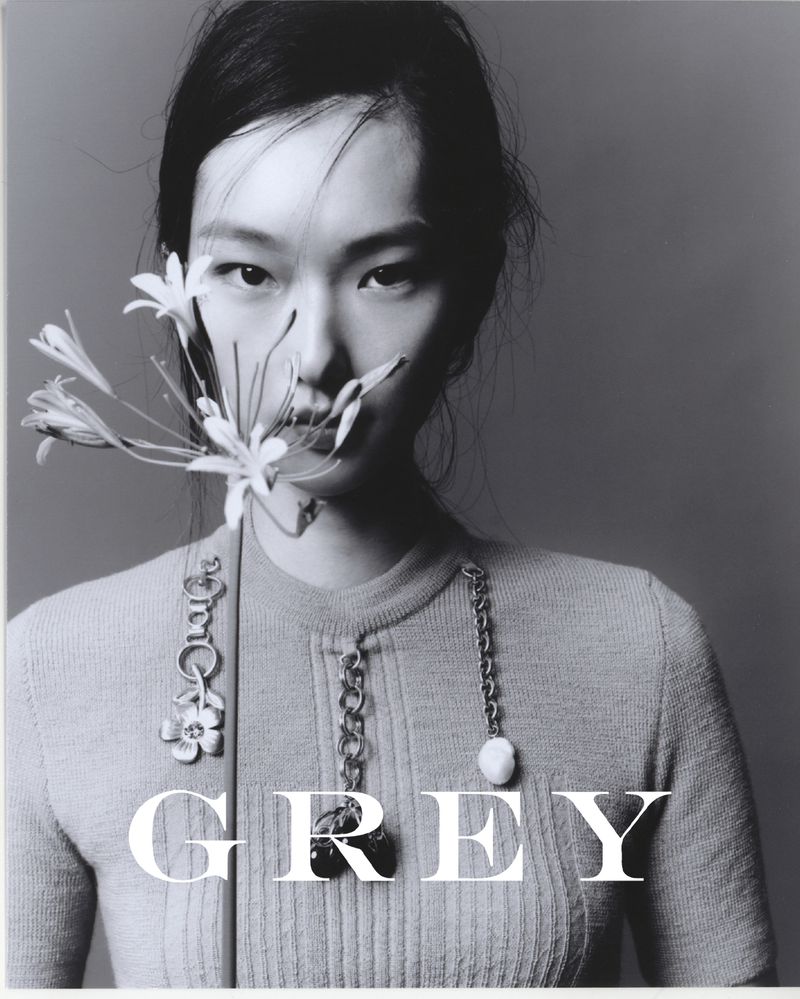 Monochromatic portrait of an Asian woman with flower, light grey background, showcasing minimalist fashion with statement necklaces.