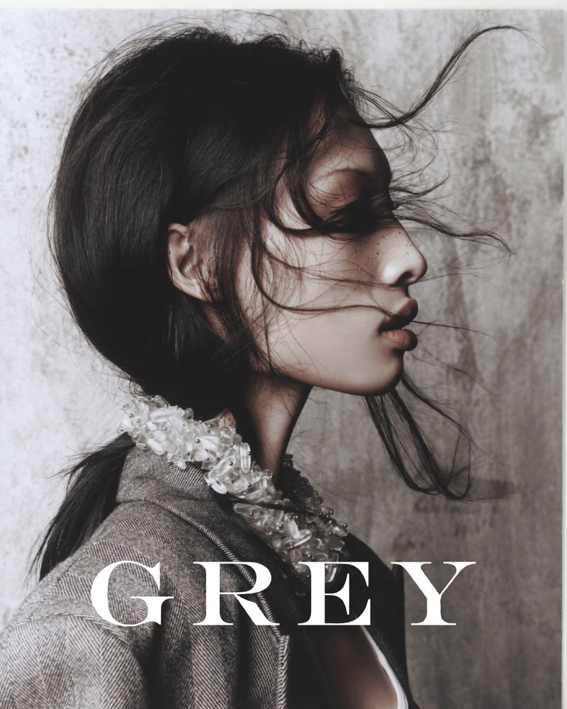Portrait of a young woman with dark hair in profile, wearing a grey jacket with decorative collar, against a neutral backdrop with prominent 'GREY' text.
