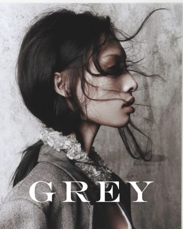 Portrait of a young woman with dark hair in profile, wearing a grey jacket with decorative collar, against a neutral backdrop with prominent 'GREY' text.