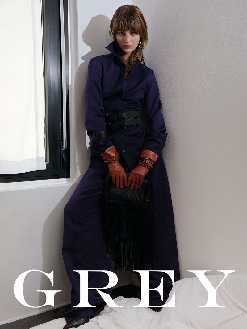 Fashion model in a blue dress and red gloves poses against a white corner with 'GREY' text overlay and a black fringed bag.