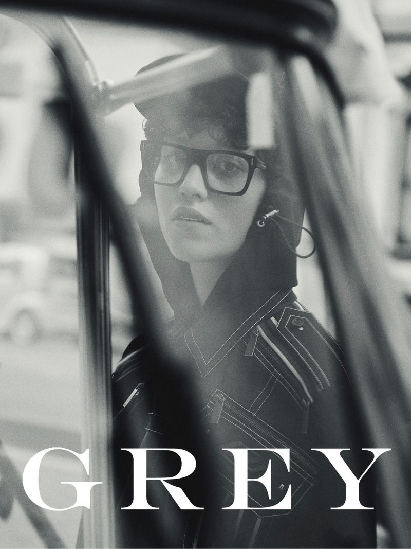 Monochrome fashion portrait of an adult female with short curly hair wearing black glasses, a hooded jacket, and dark lipstick.