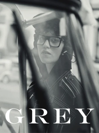 Black and white portrait of a model wearing thick black-rimmed glasses and a hooded jacket seen through a car window with GREY text.