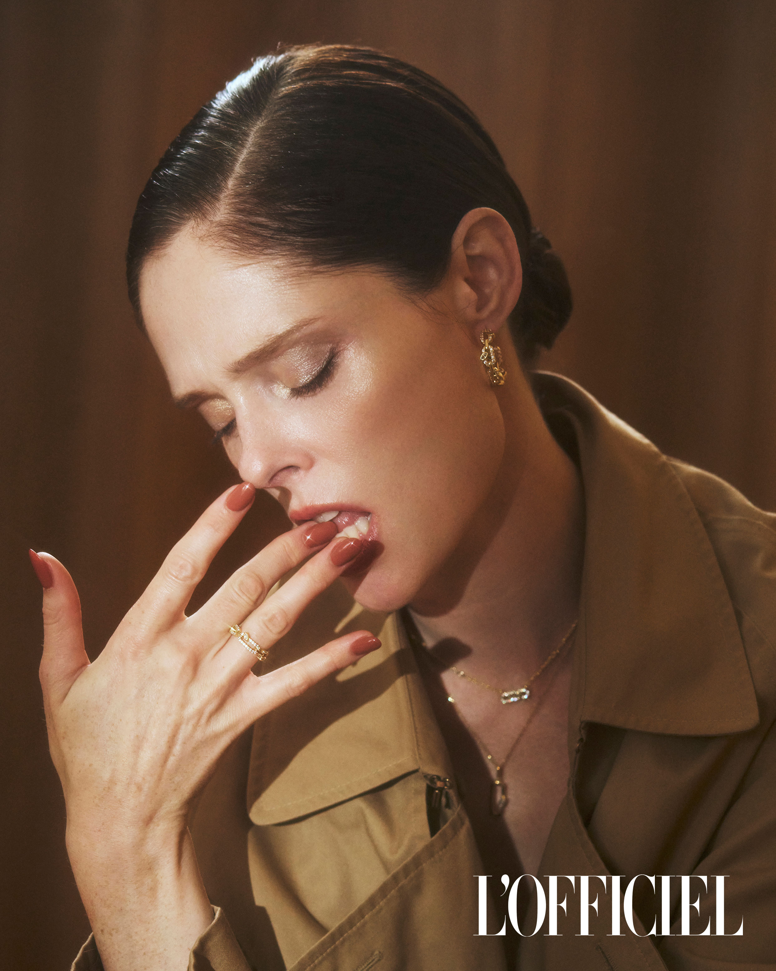 Adult woman with dark hair touches her lips, showcasing a gold ring and matching manicure; she wears gold earrings and a trench coat indoors.