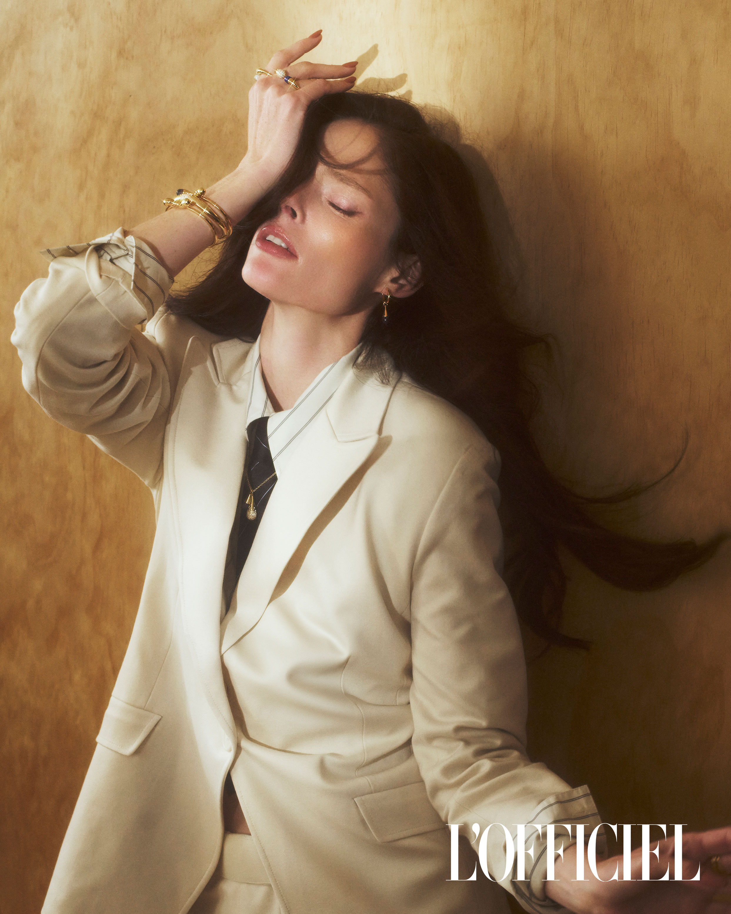 Elegant woman poses in a beige suit, accessorized with rings and bracelets, against a textured golden background, fashion magazine editorial.