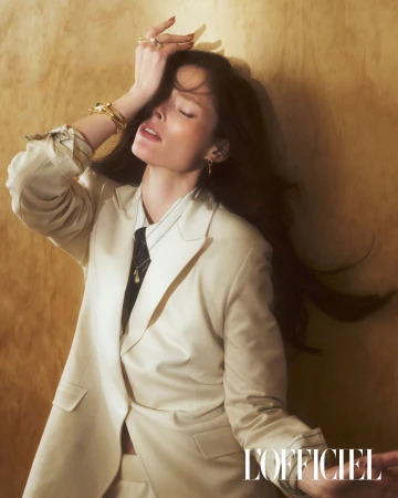 Elegant woman poses in a beige suit, accessorized with rings and bracelets, against a textured golden background, fashion magazine editorial.