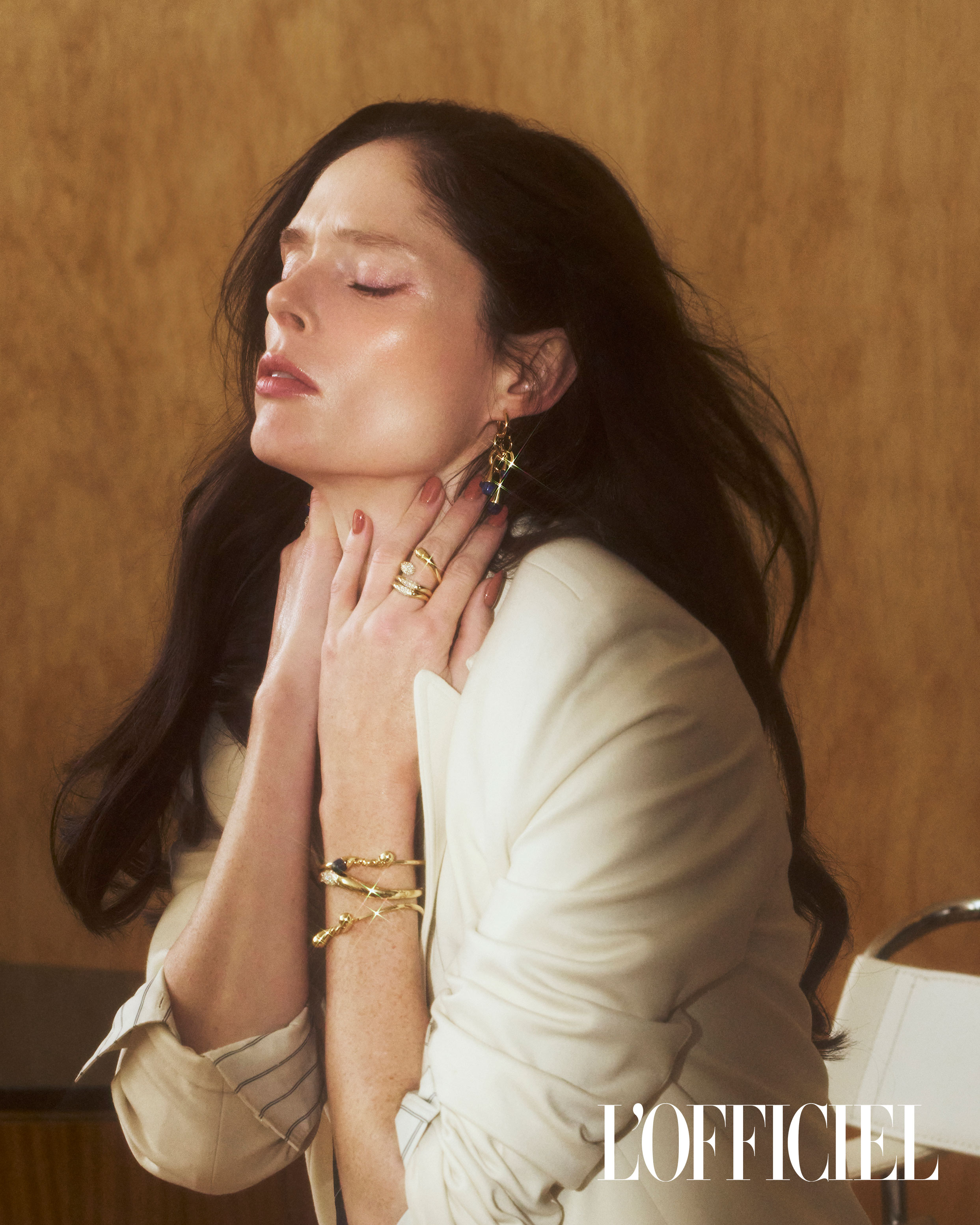 Elegant woman with dark hair wearing a white blazer and gold jewelry, posing indoors against a neutral background with sunlight.