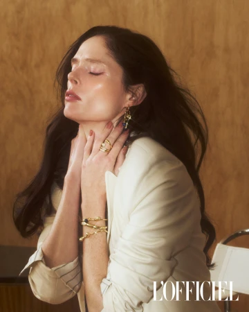 Elegant woman with dark hair wearing a white blazer and gold jewelry, posing indoors against a neutral background with sunlight.