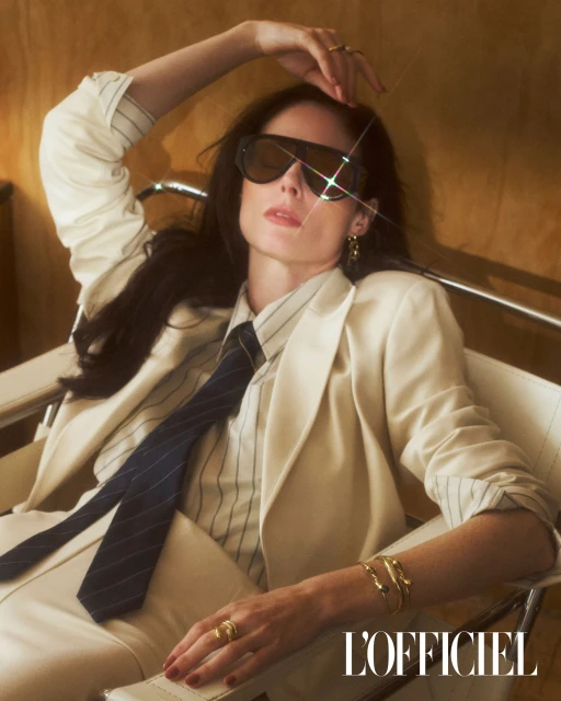 Woman in a cream suit and sunglasses poses in a chair against a brown background, wearing rings, bracelets, and a striped shirt and tie.