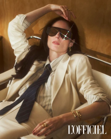 Woman in a cream suit and sunglasses poses in a chair against a brown background, wearing rings, bracelets, and a striped shirt and tie.