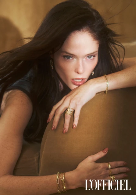 Portrait of a woman with dark hair and gold jewelry reclining in an indoor environment with warm, neutral tones and magazine logo.