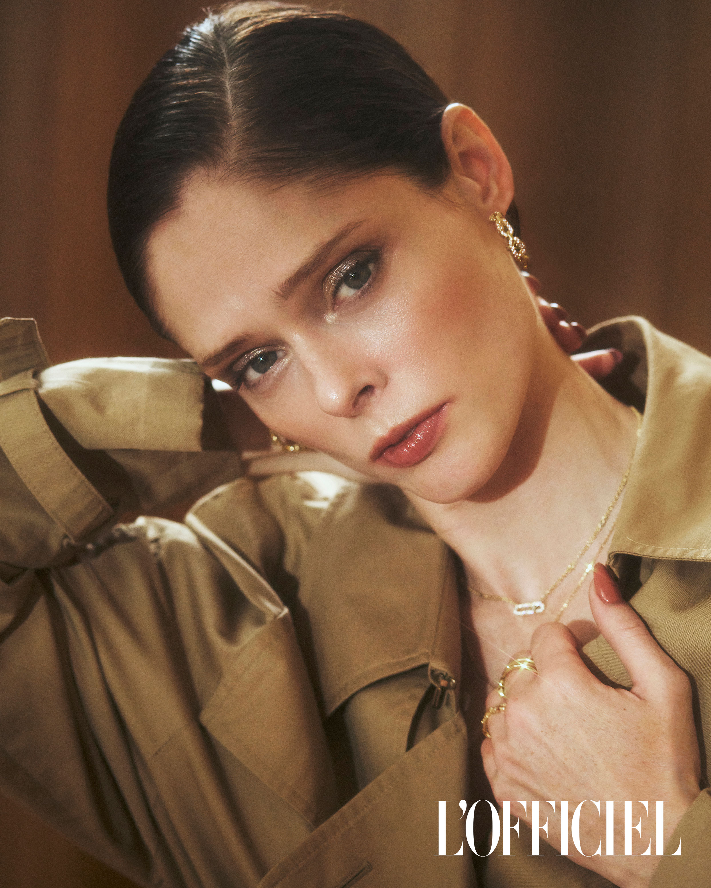 Elegant woman with gold jewelry, trench coat, and neutral makeup poses in a studio with warm lighting, showcasing a fashion editorial look.