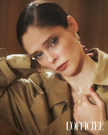 Elegant woman with gold jewelry, trench coat, and neutral makeup poses in a studio with warm lighting, showcasing a fashion editorial look.