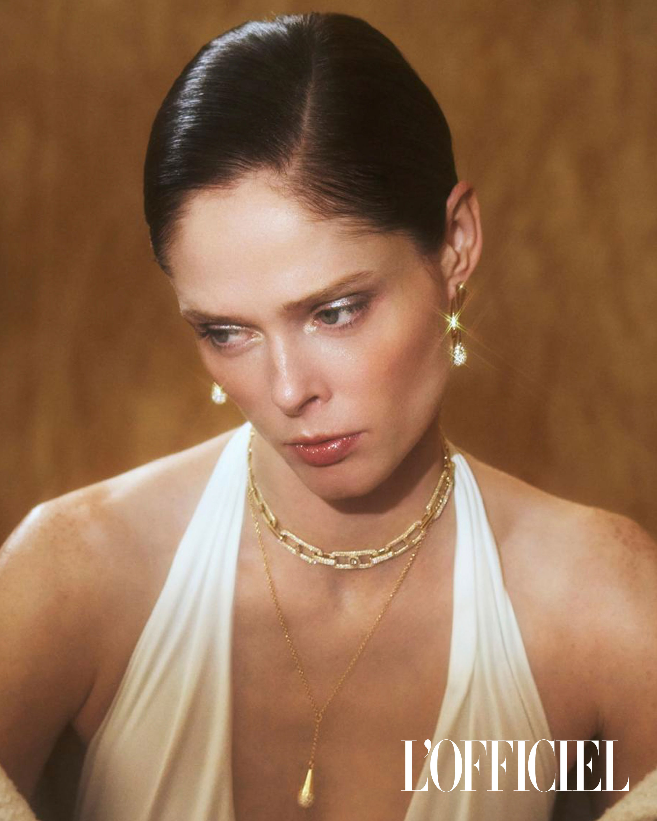 Portrait of a woman with dark hair and gold jewellery, wearing a white halter-neck top, against a warm golden background. Fashion editorial.