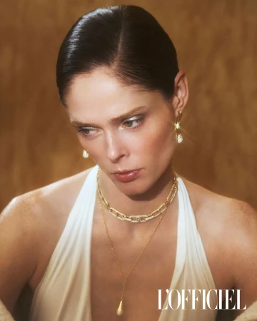 Portrait of a woman with dark hair and gold jewellery, wearing a white halter-neck top, against a warm golden background. Fashion editorial.