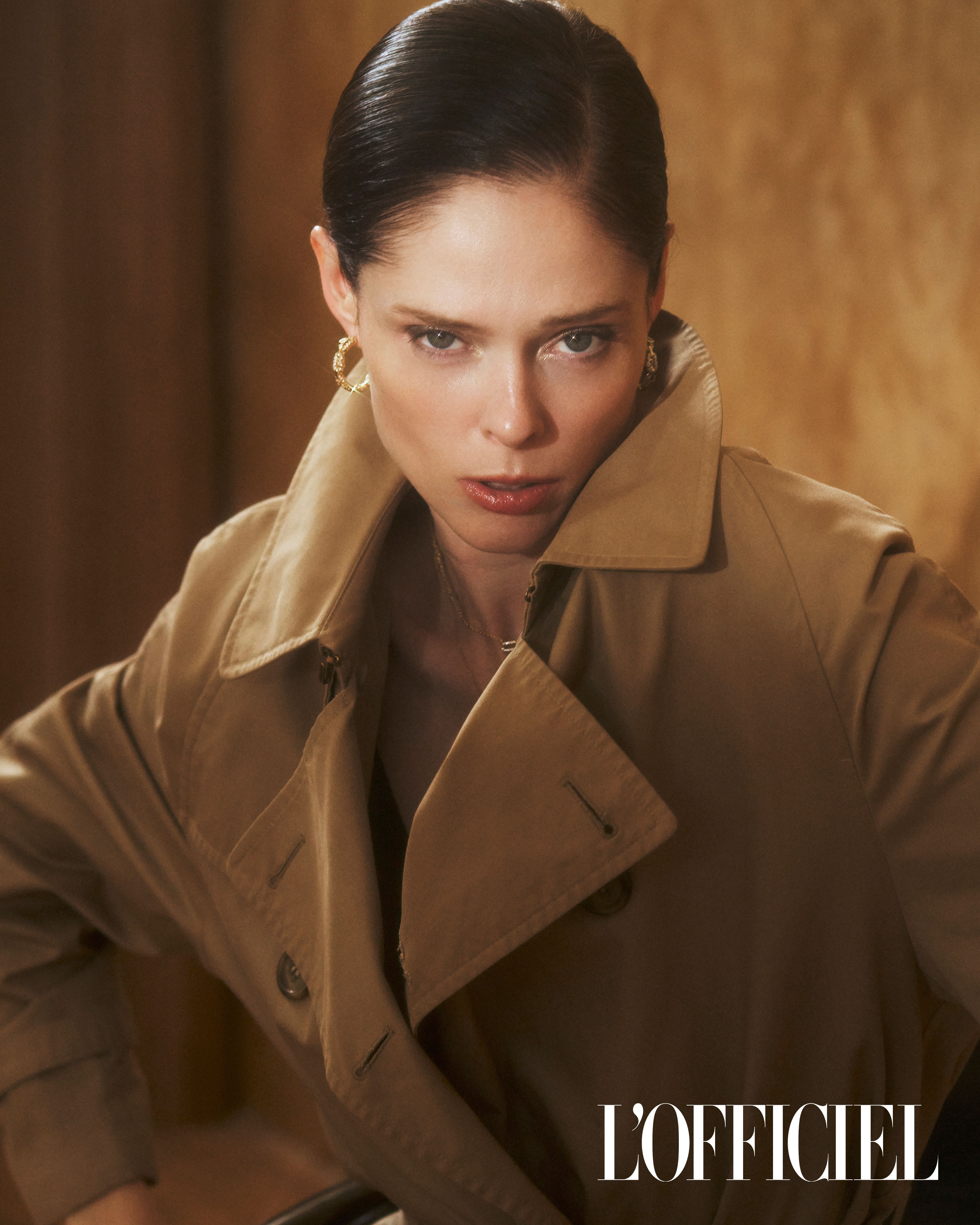 Stylish portrait of a woman in a tan trench coat with wet hair, set against a textured backdrop, captured in a studio setting.