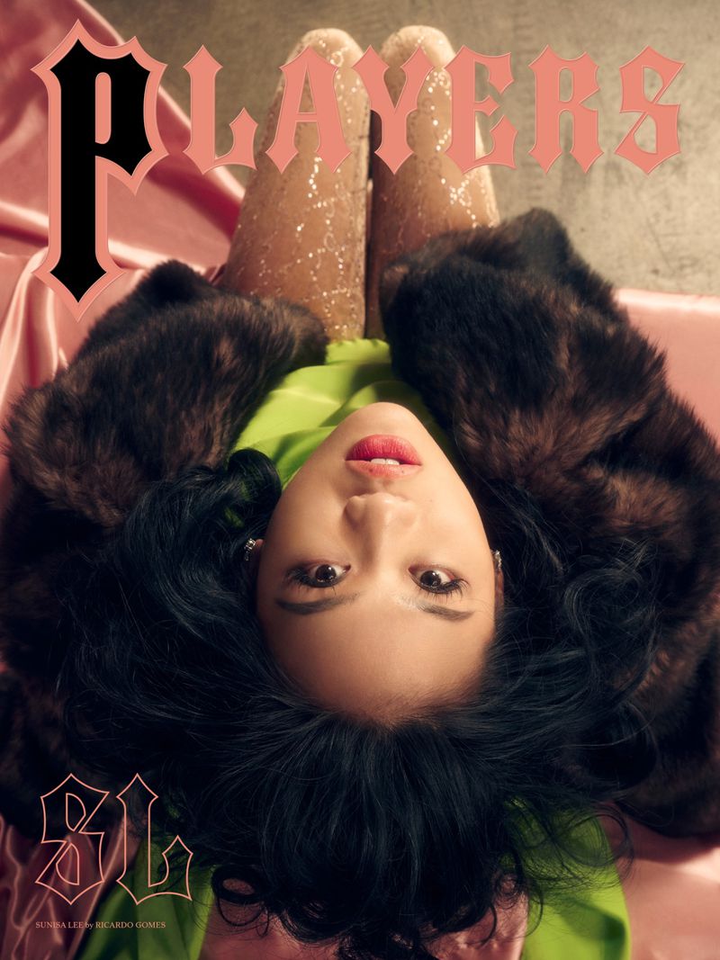 Magazine cover featuring a person with dark hair, wearing a green top and fur coat, lying on a pink satin background.
