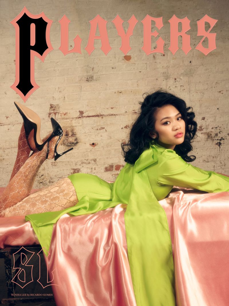 Asian woman in a lime green dress and hosiery, posing on a pink satin bed in front of a brick wall, magazine cover.