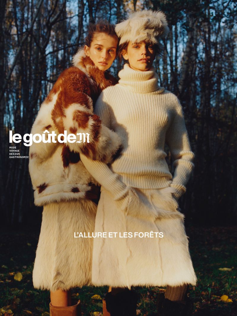 Two fashion models in fur-trimmed winter clothing pose in a forest setting with dark trees; magazine advertisement for winter fashion.