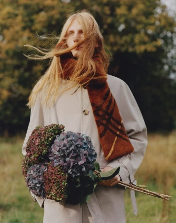 Model with windblown strawberry blonde hair in a beige trench coat and patterned rust scarf holding dark hydrangeas outdoors.
