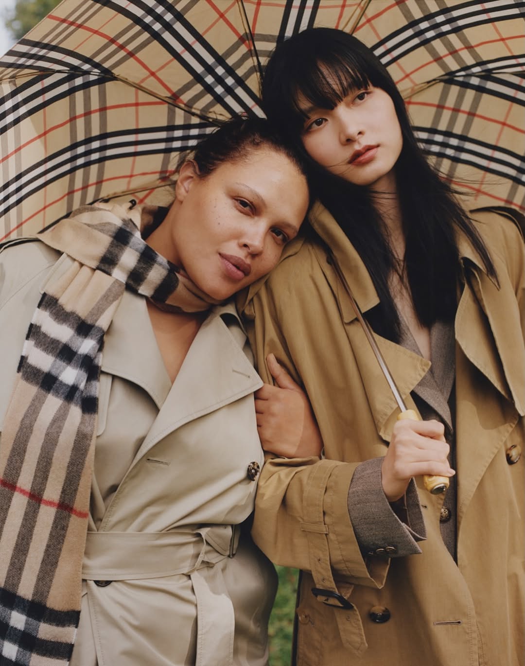 Two women in tan trench coats pose closely under a plaid umbrella and scarf, fashion, studio shot, and outdoor setting.