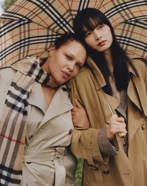 Two women in tan trench coats pose closely under a plaid umbrella and scarf, fashion, studio shot, and outdoor setting.