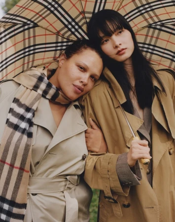 Two women in tan trench coats pose closely under a plaid umbrella and scarf, fashion, studio shot, and outdoor setting.