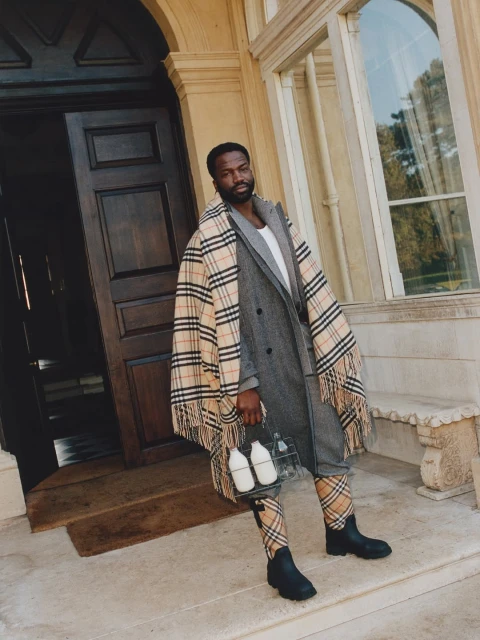 A man in a grey suit and Burberry scarf stands on the porch steps of a grand house, holding a vintage glass milk bottle carrier.