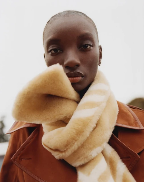 Close-up of an African-American woman in a stylish brown coat and a beige and white scarf, looking directly at the camera.
