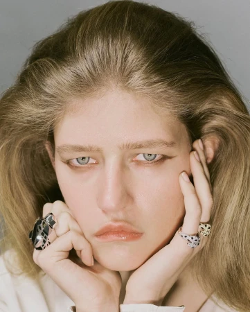 Close-up portrait of a young adult woman with blonde hair and light eyes, wearing rings and facing the camera.