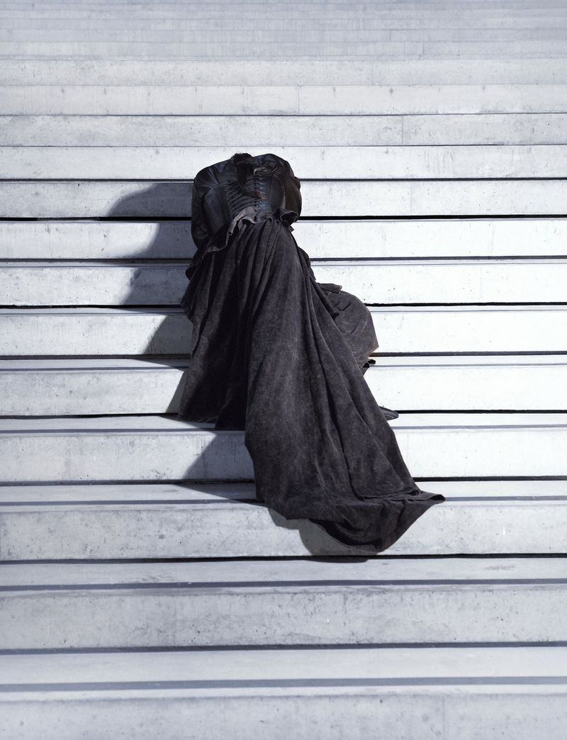 Woman in draped fabric sits on concrete stairs in a vertical shot. The fabric creates abstract shapes, and shadows define the scene.