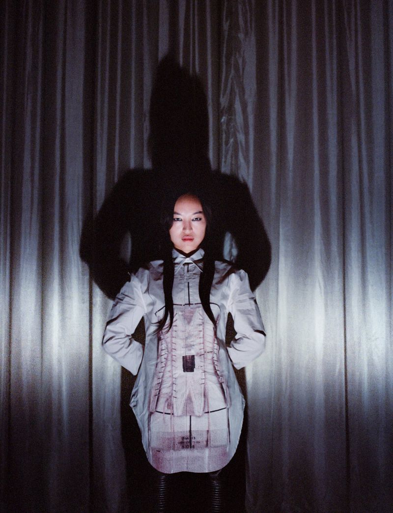 Portrait of an Asian woman in a patterned shirt with a shadow figure behind her against a curtain backdrop, creating a moody atmosphere.