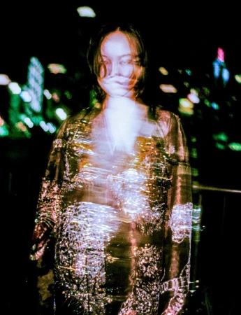 Portrait of a young woman in a shimmering top against a blurred, dark urban nightscape backdrop featuring bokeh lights.