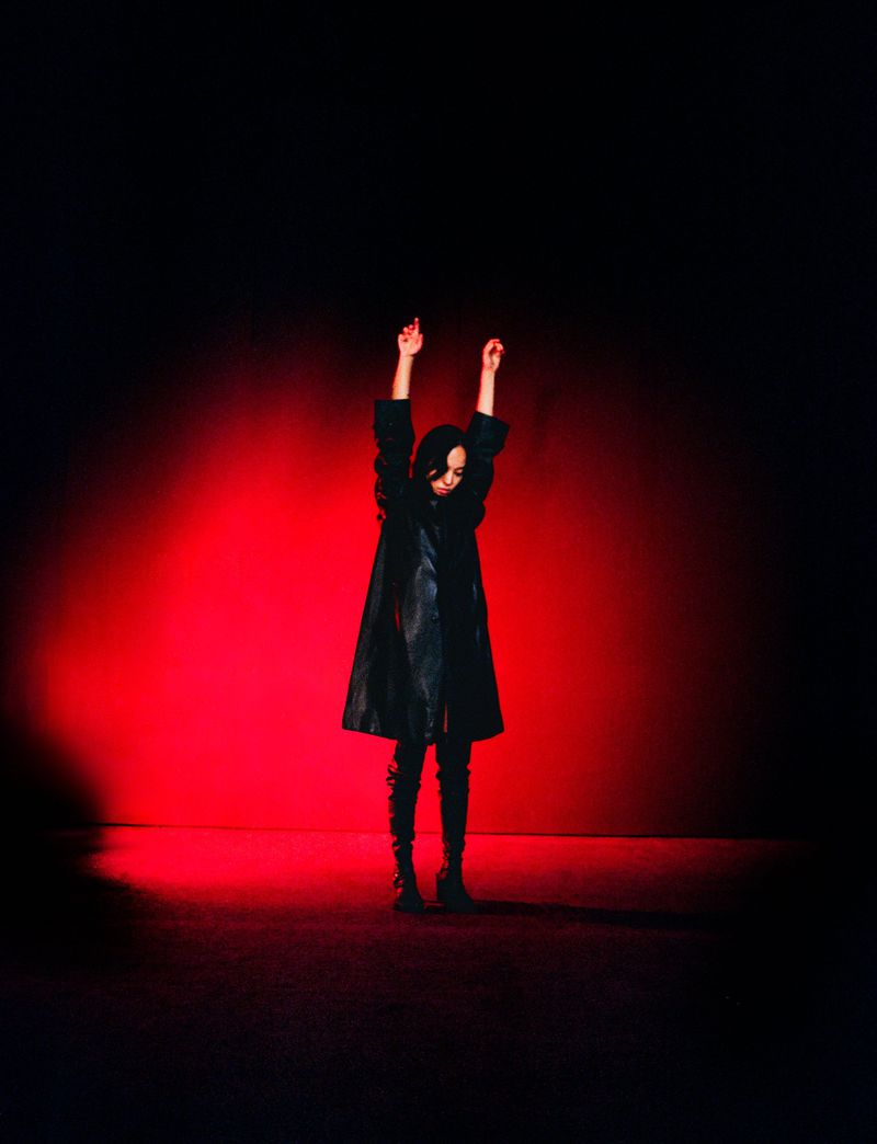 A slender figure in a black coat stands against a vibrant red and deep black backdrop, arms raised, bathed in dramatic studio lighting.