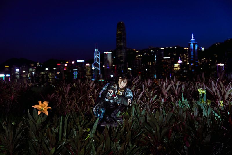 Woman in patterned jacket crouches in foliage against Hong Kong cityscape at night, captured in a medium shot with city lights.