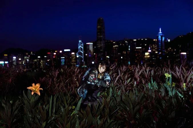 Woman in patterned jacket crouches in foliage against Hong Kong cityscape at night, captured in a medium shot with city lights.