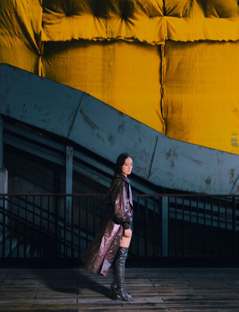 A stylish adult woman in a long, shiny coat and thigh-high boots stands on a platform against an abstract yellow and blue backdrop.