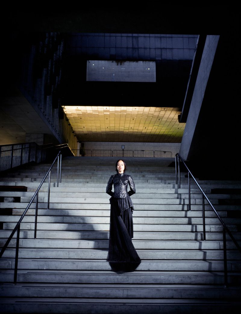 Woman in black outfit stands on concrete stairs leading toward a brightly lit tunnel, captured in a full shot with dark, shadowed tones.